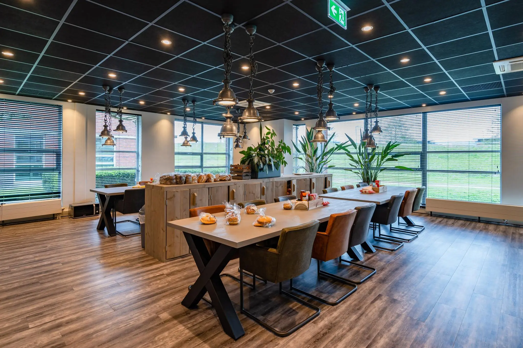 Moderne kantineruimte aan de 1e Barendrechtseweg met lange eettafels, hanglampen en verpakte broodjes.